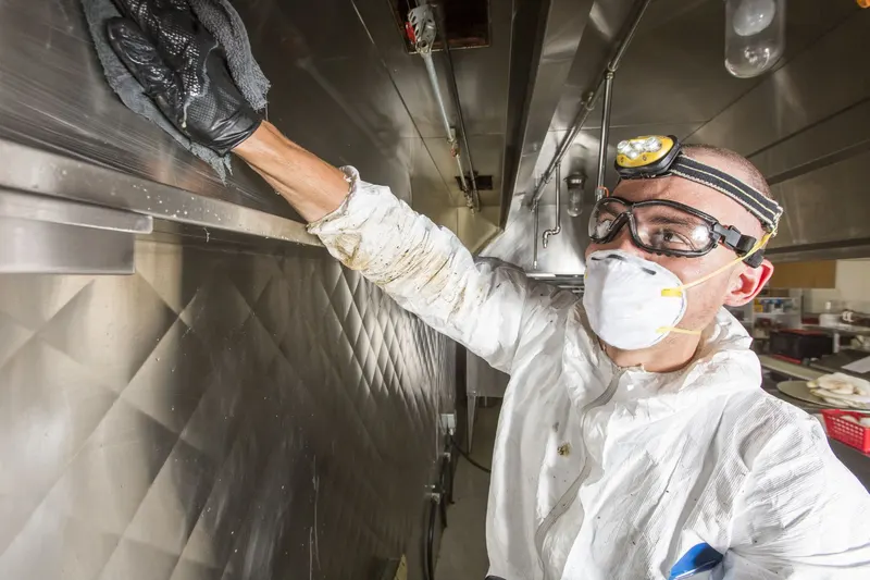 Technician in coveralls and goggles spraying degreaser onto the underside of a stainless steel commercial exhaust canopy, photographed from below