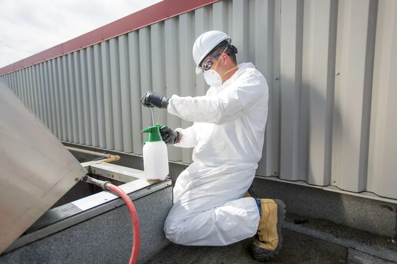 Technician in white coveralls and hard hat on a flat commercial rooftop next to an exhaust fan and a pump sprayer, low cloud overhead