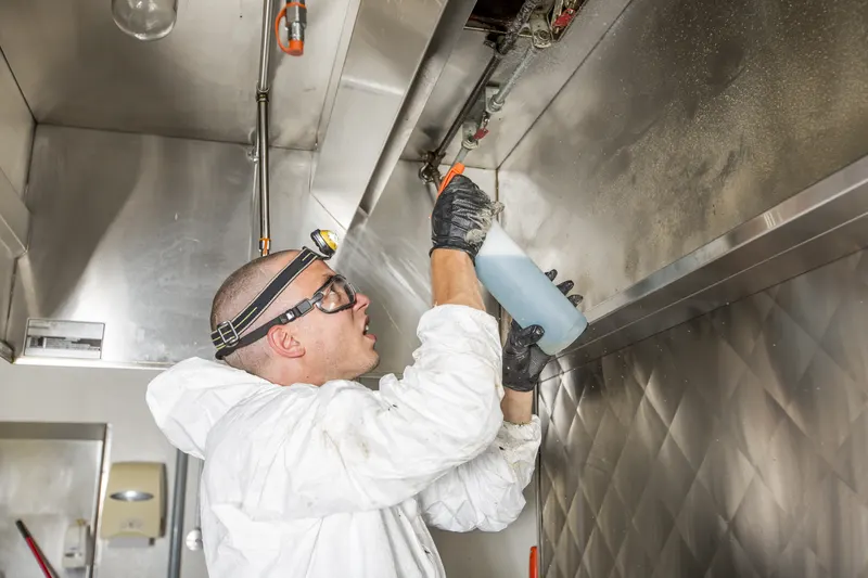 Gloved technician reaching up into the open plenum of a stainless steel commercial kitchen exhaust hood with the baffle filters removed