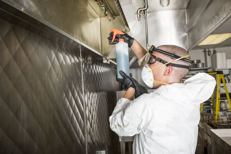 Gloved hand wiping degreaser along the bottom edge of a stainless steel kitchen exhaust canopy